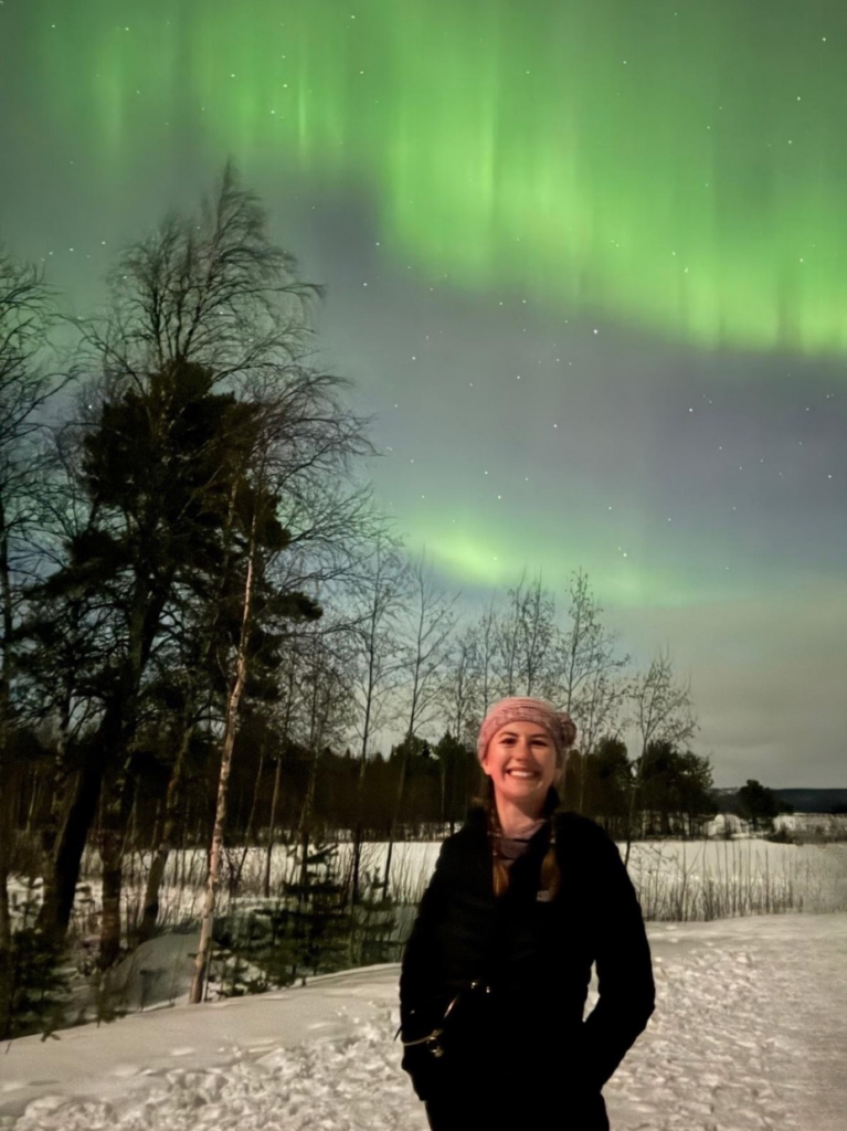 Tara Donnelly in the foreground with a snowy, forested landscape and the Northern Lights in the background