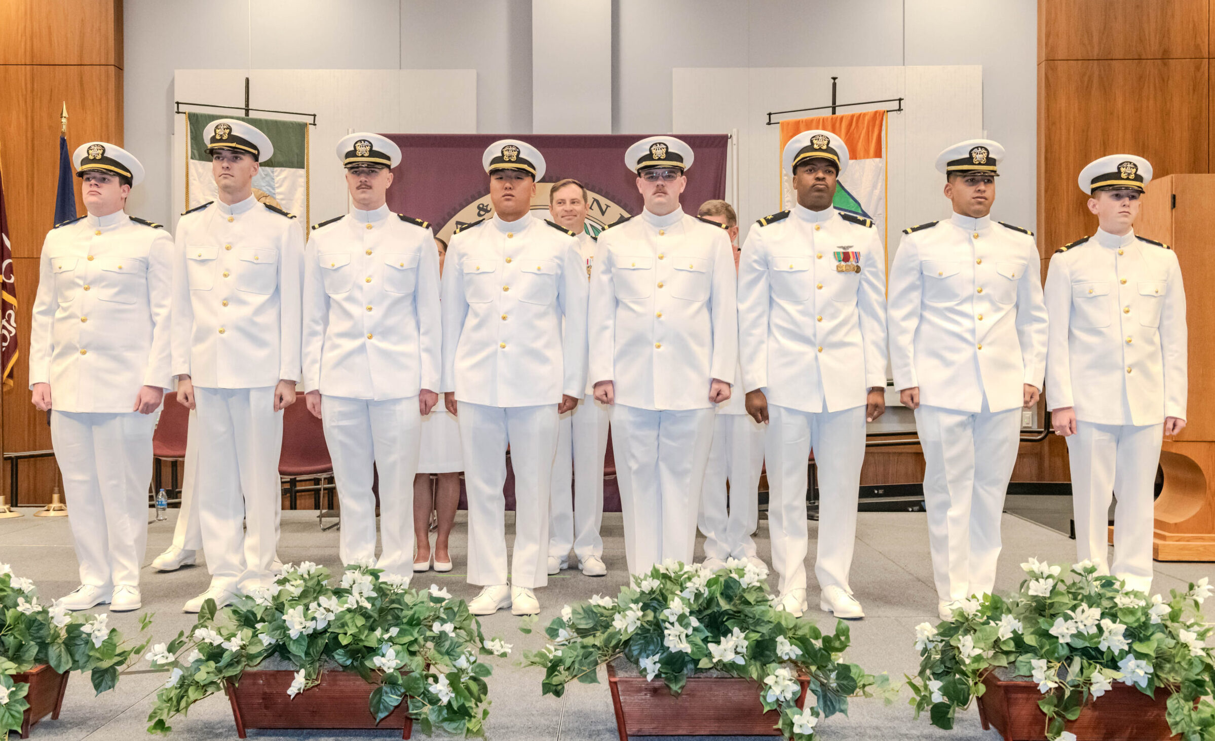 Eight new ensigns in white uniforms standing in a line on stage
