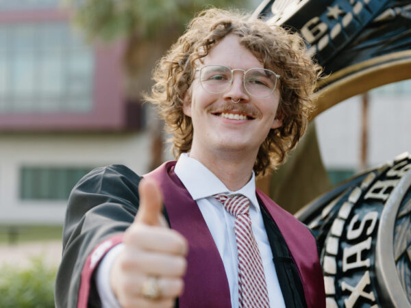 Adin Gauthier-Tyger in a "gig 'em" pose standing in front of the Aggie Ring statue at the Galveston Campus