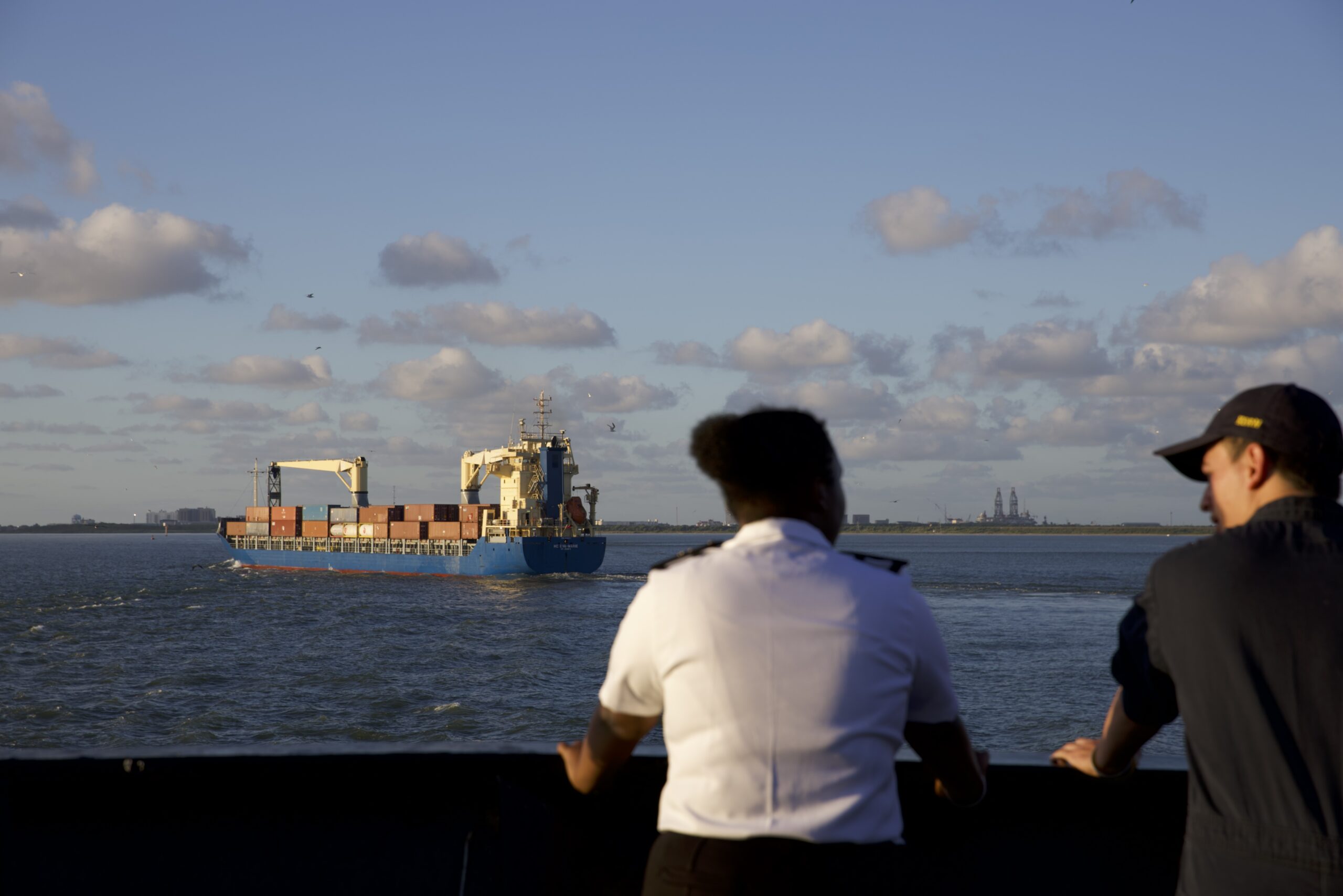 Two students, one in their salt and pepper uniform and the other in coveralls, look out over the Houston Ship Channel at a ship carrying cargo